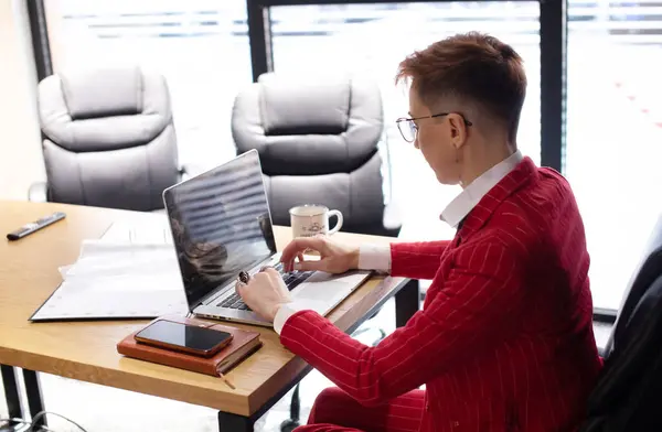 穿着红色西服的漂亮的成熟女商人的肖像和在她工作站的笔记本电脑上工作的眼镜 — 图库照片
