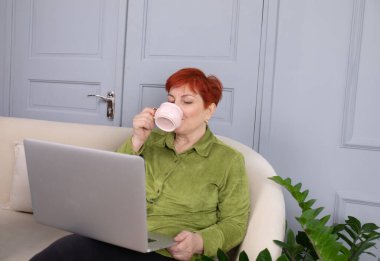 Pozitif orta yaşlı, kısa saçlı bir kadın bir fincan kahveyle kanepede oturuyor ve uzaktan bilgisayarla çalışıyor..    