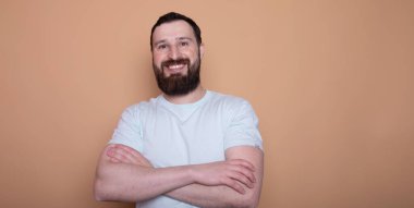 Yakışıklı sakallı genç adam kollarını kavuşturmuş ve bej arka planda izole edilmiş kameraya gülümsüyor.