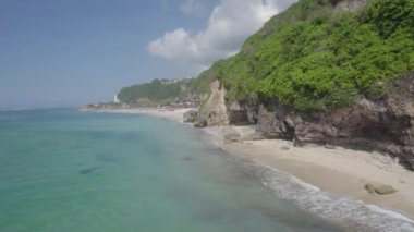 Mavi okyanusu ve arka planında yeşil ağaçları olan güzel bir sahil. Okyanus sakin ve huzurlu, gökyüzü berrak ve parlak.