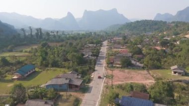 Kırmızı araba tarlada yoldan aşağı gidiyor. Yol uzun otlar, ağaçlar ve evlerle çevrili.    