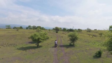 Çimenli bir arazide, toprak yolda motosiklet süren bir adamın güzel manzarası. Sumbawa, Endonezya.  