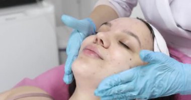 Yakın plan, mavi tıbbi eldivenli kozmetik uzmanı, cilt bakımı yapıyor, bir kadına yüz masajı yapıyor, kozmetik jeli kullanıyor, kozmetik kliniğinde ya da güzellik salonunda..   