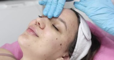 Yakın plan, mavi tıbbi eldivenli kozmetik uzmanı, cilt bakımı yapıyor, bir kadına yüz masajı yapıyor, kozmetik jeli kullanıyor, kozmetik kliniğinde ya da güzellik salonunda..   