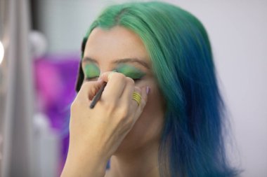 Parlak makyaj yapma süreci. Makyöz yüzüne makyaj yapan bir makyöz. Güzellik salonundaki genç yeşil saçlı kadının portresi..                         