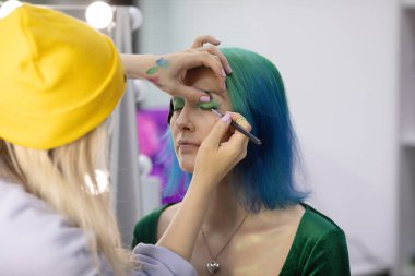 Parlak makyaj yapma süreci. Makyöz yüzüne makyaj yapan bir makyöz. Güzellik salonundaki genç yeşil saçlı kadının portresi..                         