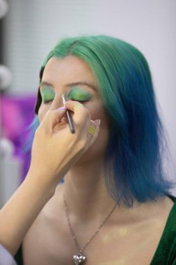 Parlak makyaj yapma süreci. Makyöz yüzüne makyaj yapan bir makyöz. Güzellik salonundaki genç yeşil saçlı kadının portresi..                         