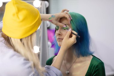 Parlak makyaj yapma süreci. Makyöz yüzüne makyaj yapan bir makyöz. Güzellik salonundaki genç yeşil saçlı kadının portresi..                         