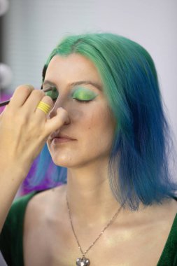 Parlak makyaj yapma süreci. Makyöz yüzüne makyaj yapan bir makyöz. Güzellik salonundaki genç yeşil saçlı kadının portresi..                         
