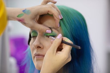 Parlak makyaj yapma süreci. Makyöz yüzüne makyaj yapan bir makyöz. Güzellik salonundaki genç yeşil saçlı kadının portresi..                         