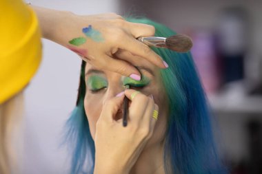 Parlak makyaj yapma süreci. Makyöz yüzüne makyaj yapan bir makyöz. Güzellik salonundaki genç yeşil saçlı kadının portresi..                         