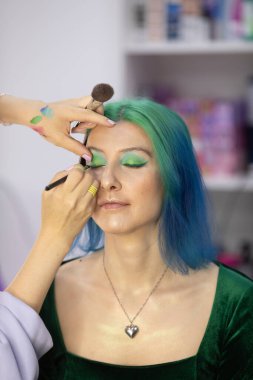Parlak makyaj yapma süreci. Makyöz yüzüne makyaj yapan bir makyöz. Güzellik salonundaki genç yeşil saçlı kadının portresi..                         
