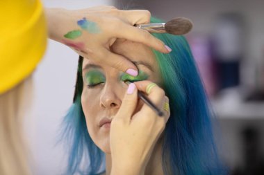 Parlak makyaj yapma süreci. Makyöz yüzüne makyaj yapan bir makyöz. Güzellik salonundaki genç yeşil saçlı kadının portresi..                         