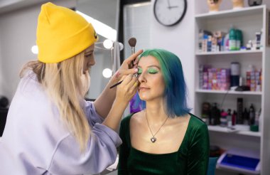 Parlak makyaj yapma süreci. Makyöz yüzüne makyaj yapan bir makyöz. Güzellik salonundaki genç yeşil saçlı kadının portresi..                         