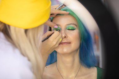 Parlak makyaj yapma süreci. Makyöz yüzüne makyaj yapan bir makyöz. Güzellik salonundaki genç yeşil saçlı kadının portresi..                         