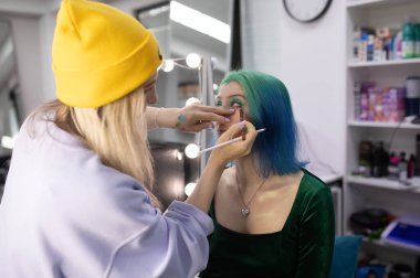 Parlak makyaj yapma süreci. Makyöz yüzüne makyaj yapan bir makyöz. Güzellik salonundaki genç yeşil saçlı kadının portresi..                         