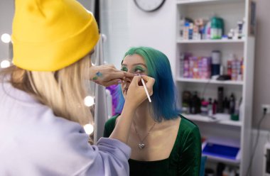 Parlak makyaj yapma süreci. Makyöz yüzüne makyaj yapan bir makyöz. Güzellik salonundaki genç yeşil saçlı kadının portresi..                         