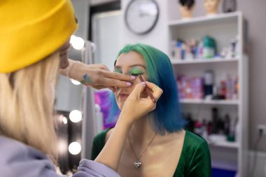 Parlak makyaj yapma süreci. Makyöz yüzüne makyaj yapan bir makyöz. Güzellik salonundaki genç yeşil saçlı kadının portresi..                         