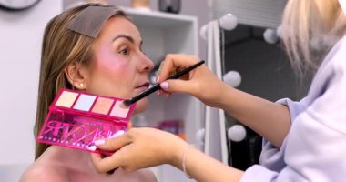 Makyöz, güzellik salonunda genç bir kadının profesyonel makyajını yapıyor.                