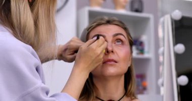 Makyöz, güzellik salonunda genç bir kadının profesyonel makyajını yapıyor.                