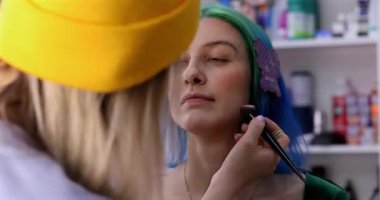 Parlak makyaj yapma süreci. Makyöz yüzüne makyaj yapan bir makyöz. Güzellik salonundaki genç yeşil saçlı kadının portresi..                        