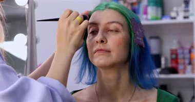 Parlak makyaj yapma süreci. Makyöz yüzüne makyaj yapan bir makyöz. Güzellik salonundaki genç yeşil saçlı kadının portresi..                        