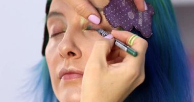 Parlak makyaj yapma süreci. Makyöz yüzüne makyaj yapan bir makyöz. Güzellik salonundaki genç yeşil saçlı kadının portresi..                        