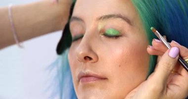 Parlak makyaj yapma süreci. Makyöz yüzüne makyaj yapan bir makyöz. Güzellik salonundaki genç yeşil saçlı kadının portresi..                        