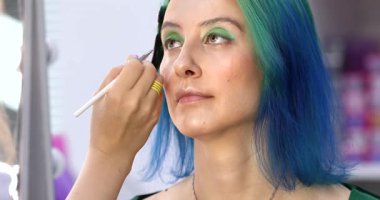 Parlak makyaj yapma süreci. Makyöz yüzüne makyaj yapan bir makyöz. Güzellik salonundaki genç yeşil saçlı kadının portresi..                        