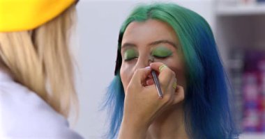 Parlak makyaj yapma süreci. Makyöz yüzüne makyaj yapan bir makyöz. Güzellik salonundaki genç yeşil saçlı kadının portresi..                        