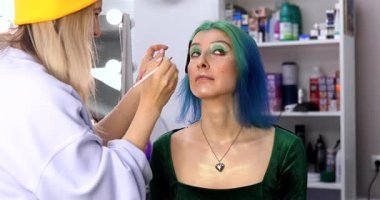 Parlak makyaj yapma süreci. Makyöz yüzüne makyaj yapan bir makyöz. Güzellik salonundaki genç yeşil saçlı kadının portresi..                        