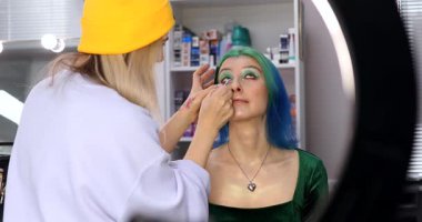 Parlak makyaj yapma süreci. Makyöz yüzüne makyaj yapan bir makyöz. Güzellik salonundaki genç yeşil saçlı kadının portresi..                        