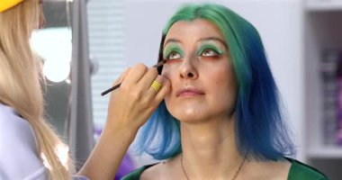 Parlak makyaj yapma süreci. Makyöz yüzüne makyaj yapan bir makyöz. Güzellik salonundaki genç yeşil saçlı kadının portresi..                        