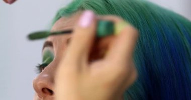 Parlak makyaj yapma süreci. Makyöz yüzüne makyaj yapan bir makyöz. Güzellik salonundaki genç yeşil saçlı kadının portresi..                        