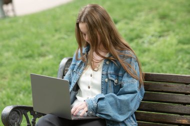 Kot ceketli genç kadın bankta oturuyor ve dizüstü bilgisayar kullanıyor..        
