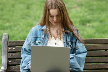 Kot ceketli genç kadın bankta oturuyor ve dizüstü bilgisayar kullanıyor..        