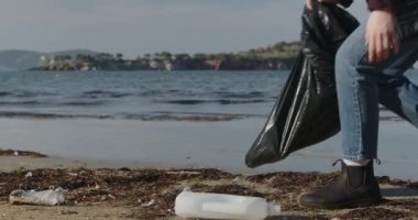 eco-activist girl collects plastic trash on the coast. Young woman collects plastic garbage in a garbage bag on the sandy beach of the sea. Girl cleans ocean coastline from plastic bottle trash on