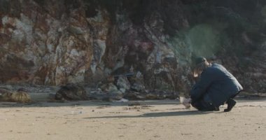 Çevreci kız sahilde plastik çöp topluyor. Genç bir kadın denizin kumlu sahilinde çöp torbasında plastik çöp topluyor. Kız, okyanus kıyısını plastik şişe çöpünden temizliyor.