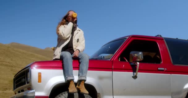 迷人的长发女孩站在山景背景下的老式汽车旁边 喝着热杯子里的饮料 高质量的4K镜头 — 图库视频影像