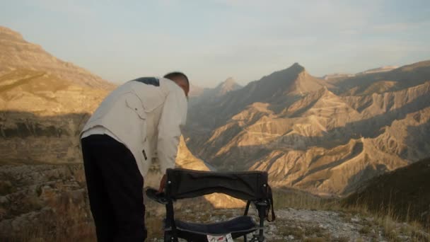 若い魅力的なヒップスター男性は 山の乾燥した南部の風景を背景にクリアリングでキャンプ折りたたみ椅子に座って 距離を調べる 高品質4K映像 — ストック動画