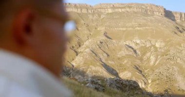 Genç ve çekici hippi bir adam, dağlık kuru bir güney manzarasının zeminine karşı bir açıklıkta kamp sandalyesinde oturuyor ve uzaklığa bakıyor. Yüksek kalite 4k görüntü