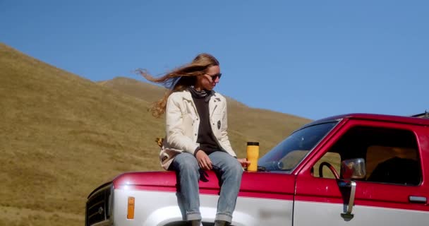 迷人的长发女孩站在山景背景下的老式汽车旁边 喝着热杯子里的饮料 高质量的4K镜头 — 图库视频影像
