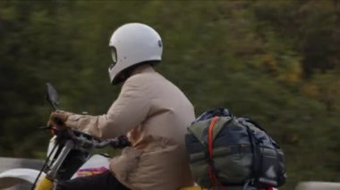 Bej ceketli bir motosikletçi, kemeriyle bağlanmış bohçalarla bir yolculuğa çıkar, macera duygusunu pratiklikle harmanlar. Sahne klasik ve off-road motosikletlerinin karışımı ile açılır.