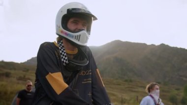 Çarpıcı bir dağ manzarasının ortasında, genç bir adam eski bir off-road motosikletine binmiş, maceraperest ruhunu gösteren beyaz bir miğfer takıyor. Açık yüzlü miğfer rüzgarın