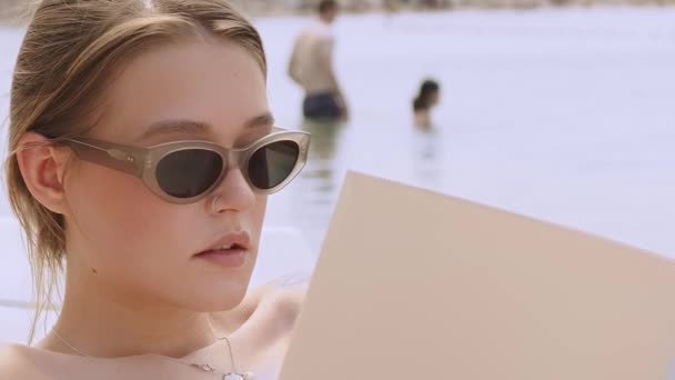 一个戴着太阳镜的女孩在海滩上的画像 一位年轻漂亮的姑娘躺在海滩上或游泳池边看书或杂志 在后台 一个男人和一个女孩在海里或游泳池里玩得很开心 — 图库视频影像