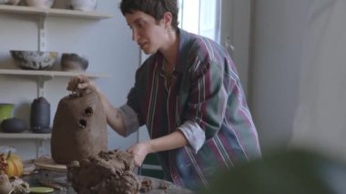 Sanatsal ifadenin keşfedilmemiş diyarlarında büyüleyici bir yolculuğa çıkın. Büyüleyici kısa saçlı bir güzellik, erkeksi bir gömlek giymiş, tutkulu bir şekilde kilden bir vazo yontuyor.