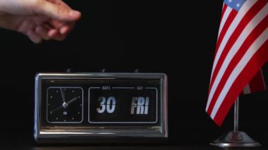 El, arka planda küçük bir Amerikan bayrağı bulunan bir ters saat üzerindeki tarihi ayarlıyor. Amerika 'daki zaman yönetimi ve teslim tarihlerini sembolize ediyor. Vintage kapaklı saatin elle ayarlanması
