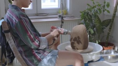 Kil vazoyu kurutan, saç kurutma makinesiyle kurutan ve sandalyede oturan kız seramik sanatçısı. Vazo kil pervazda dönüyor. Videosu yaratıcı sürecin kutlaması.