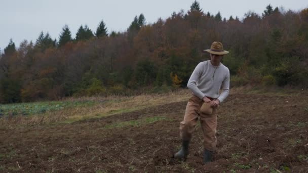 播种收获的种子 农民穿着乡村服装步行穿过秋天的天空下的田野 高质量的4K镜头 作为一个农民 穿着乡村服装 头戴帽子 踏上乡村风情之旅 — 图库视频影像