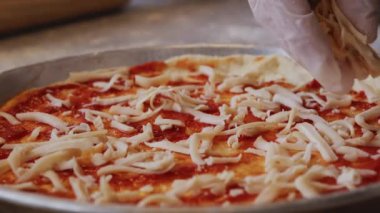 Önlüklü bir adam, un serpiştirilmiş bir masaya pizza hamuru tabanı üzerine domates ezmesi sürüyor. Önündeki masada pizza malzemeleriyle dolu kaseler var. Hamurun kenarlarını çimdikliyor.